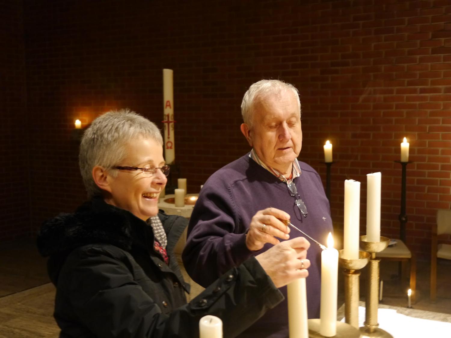 Eine Küsterin und ein Küster zünden gemeinsam Kerzen in einer Kirche an.